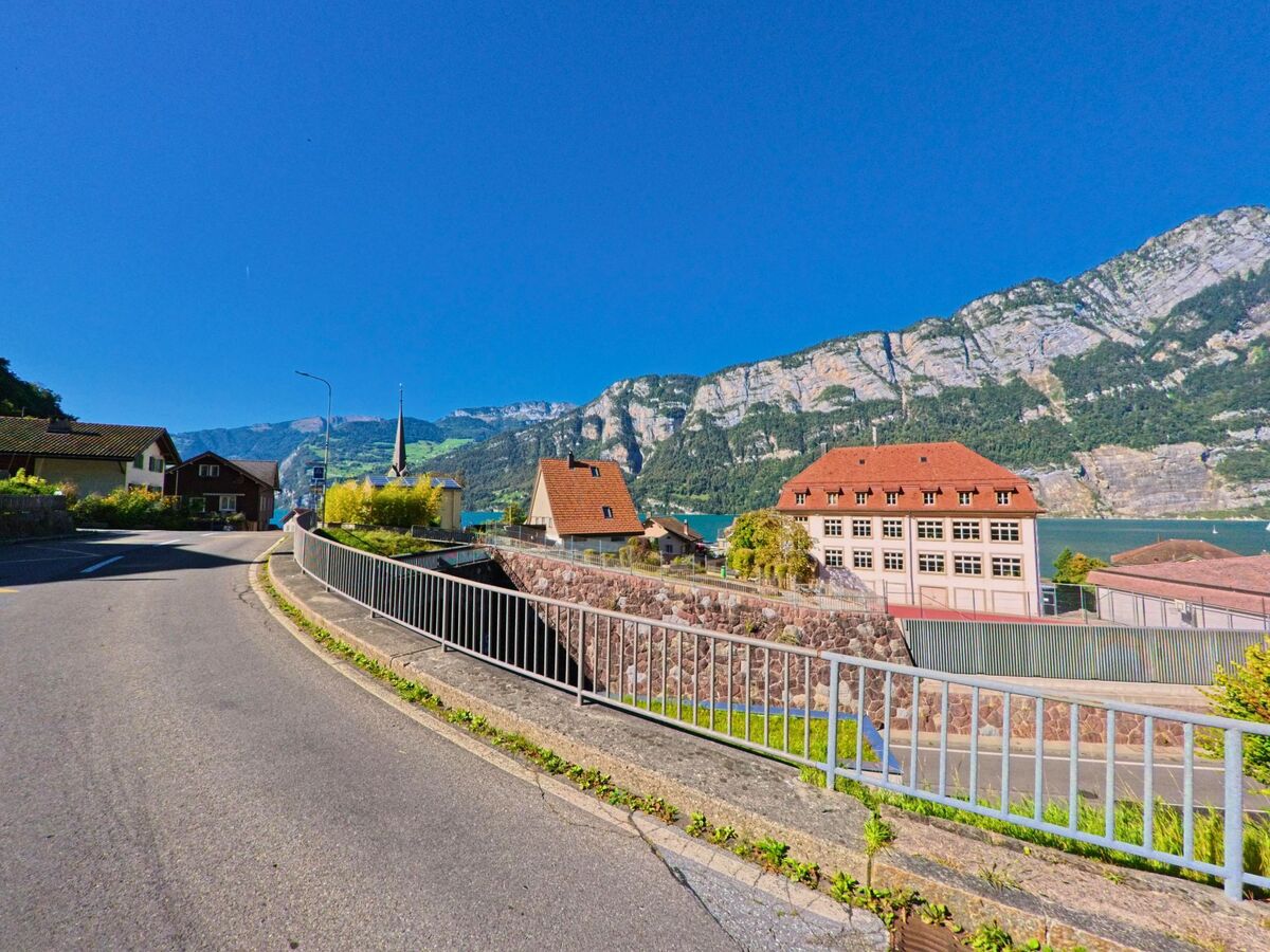 Entlang einer leicht ansteigenden Strasse mit Geländer öffnet sich der Blick auf Wohnhäuser und ein markantes Gebäude vor steilen Felswänden in Glarus Nord (8865, Kanton Glarus).