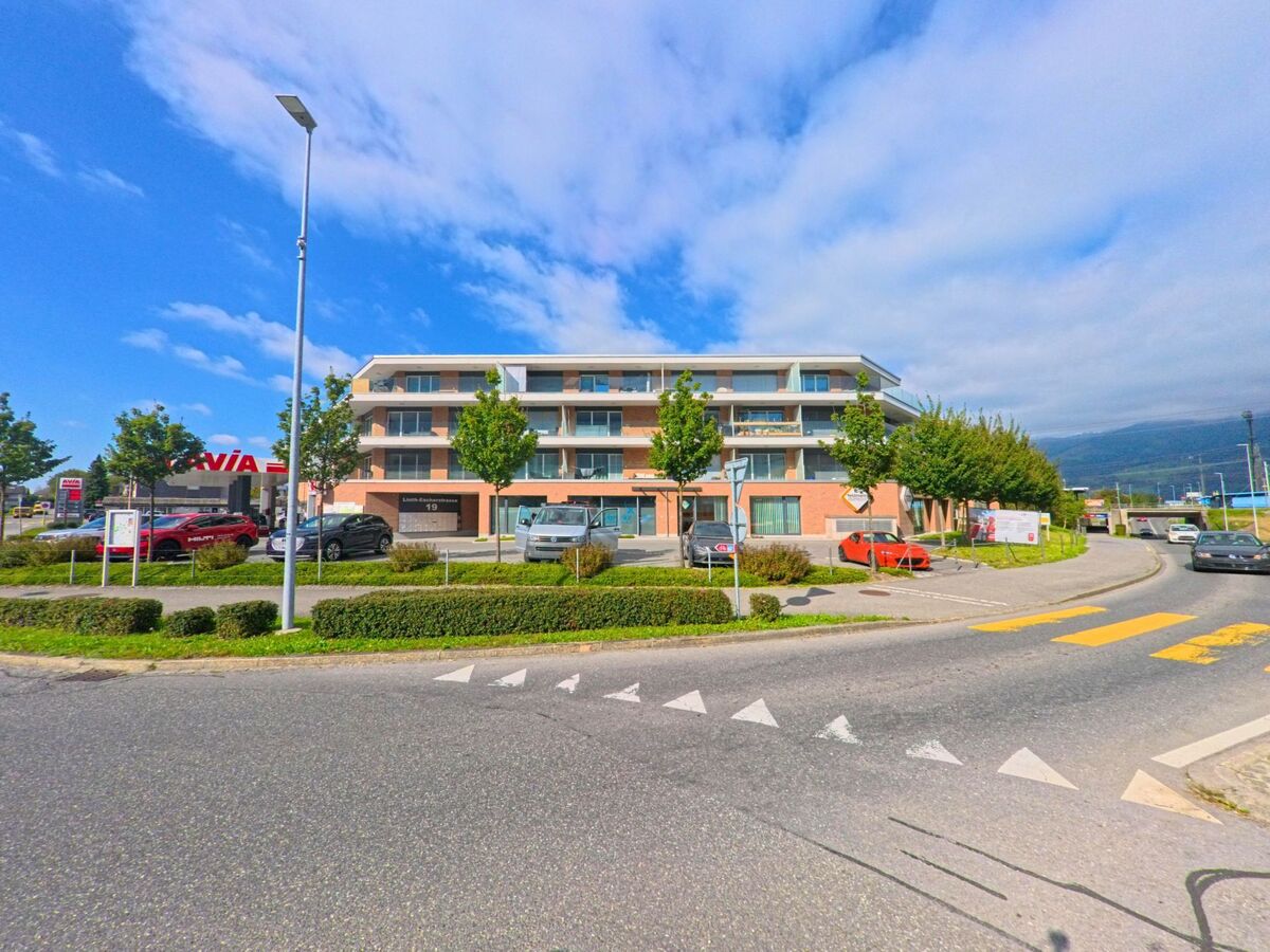 Entlang einer breiten Strasse mit Markierungen und Bepflanzung steht ein modernes Wohn- und Geschäftsgebäude in Glarus Nord (8865, Kanton Glarus), während sich dahinter die umliegende Landschaft öffnet.
