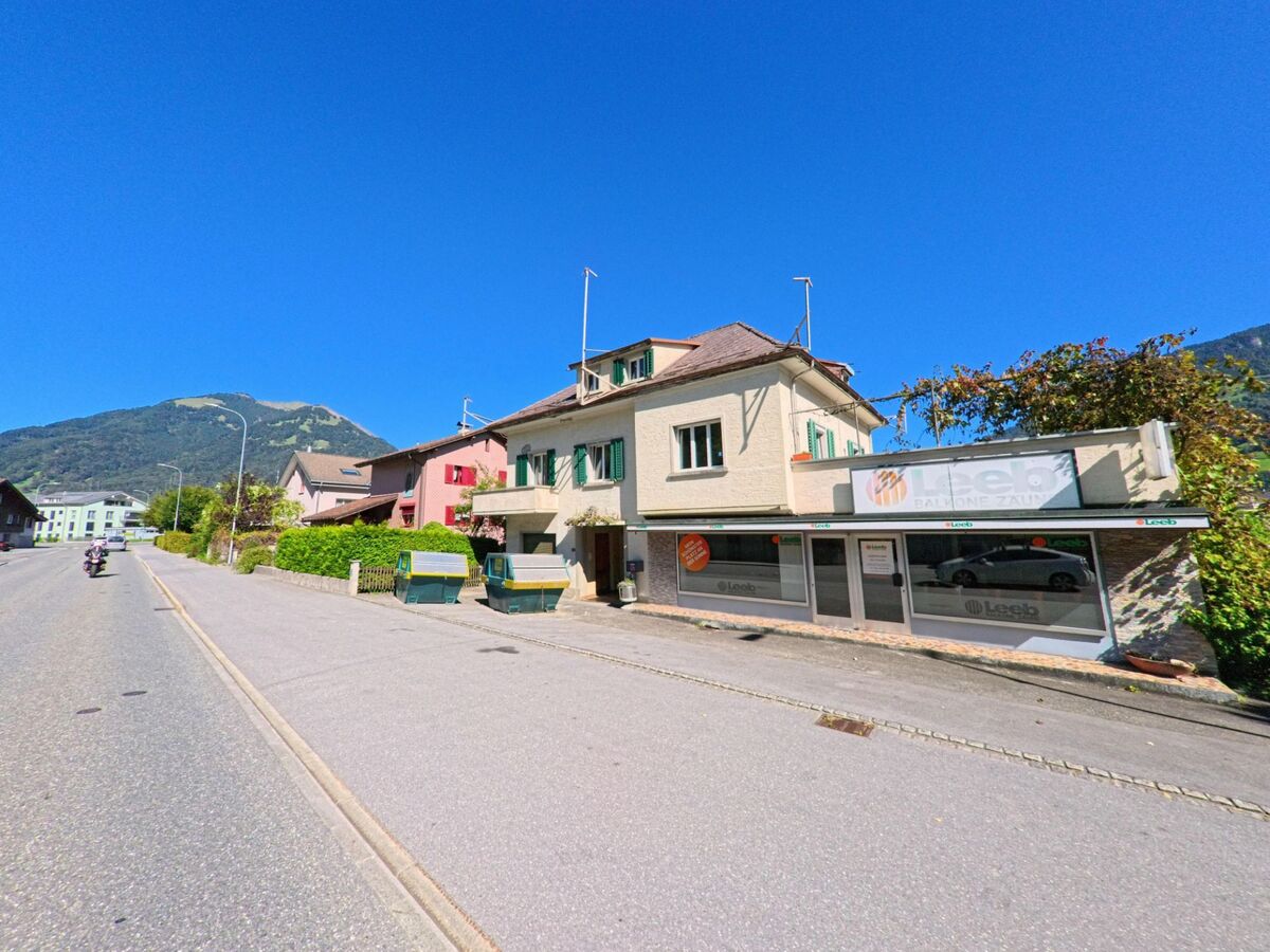 Entlang einer breiten Strasse reihen sich Wohn- und Gewerbebauten mit Schaufenstern in Glarus Nord (8865, Kanton Glarus), wobei im Hintergrund die umliegenden Hügel sichtbar sind.