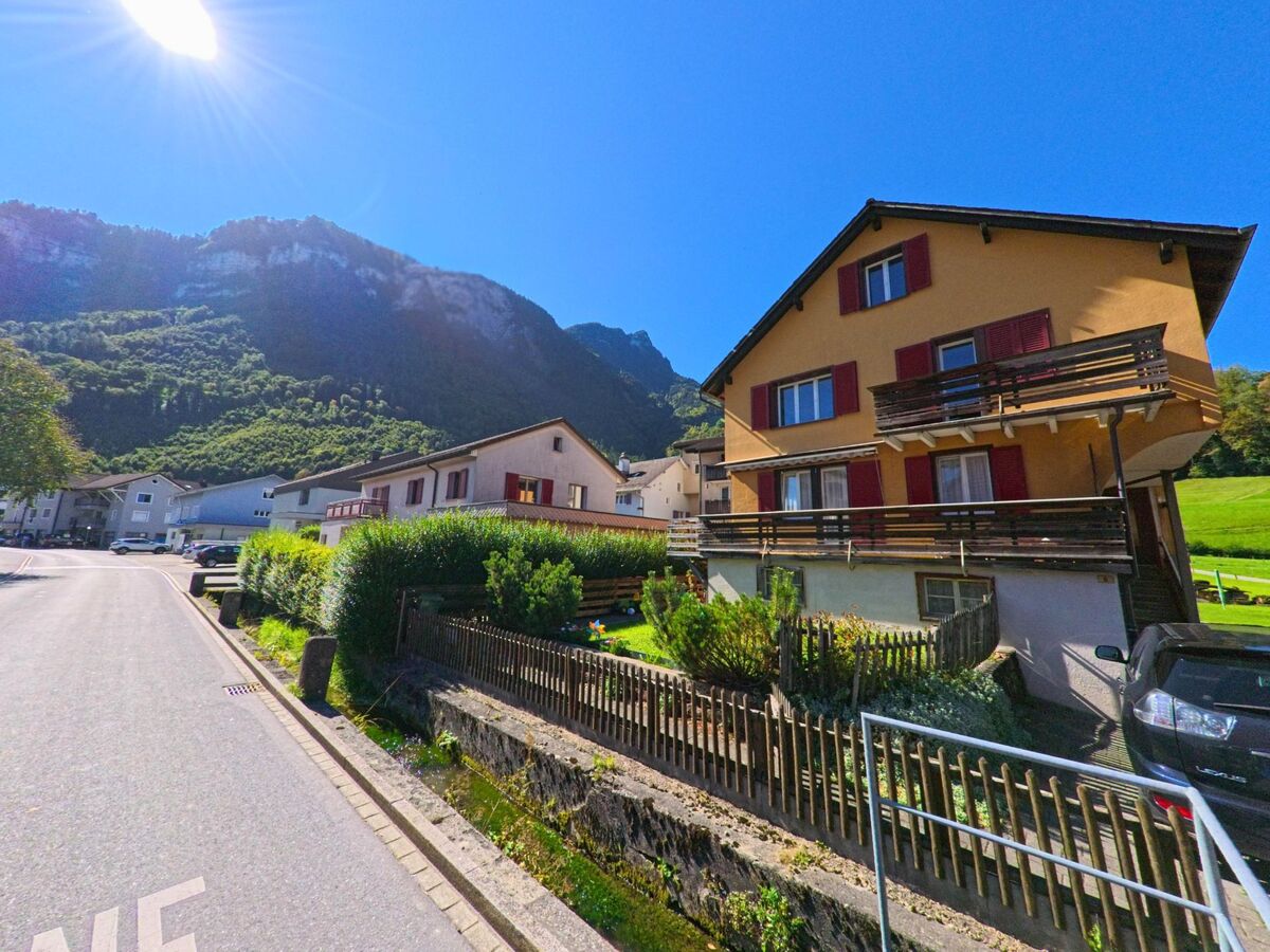 Entlang einer ruhigen Quartierstrasse stehen Wohnhäuser mit Balkonen und Gärten in Glarus Nord (8865, Kanton Glarus), während sich im Hintergrund bewaldete Berghänge unter klarem Himmel erheben.