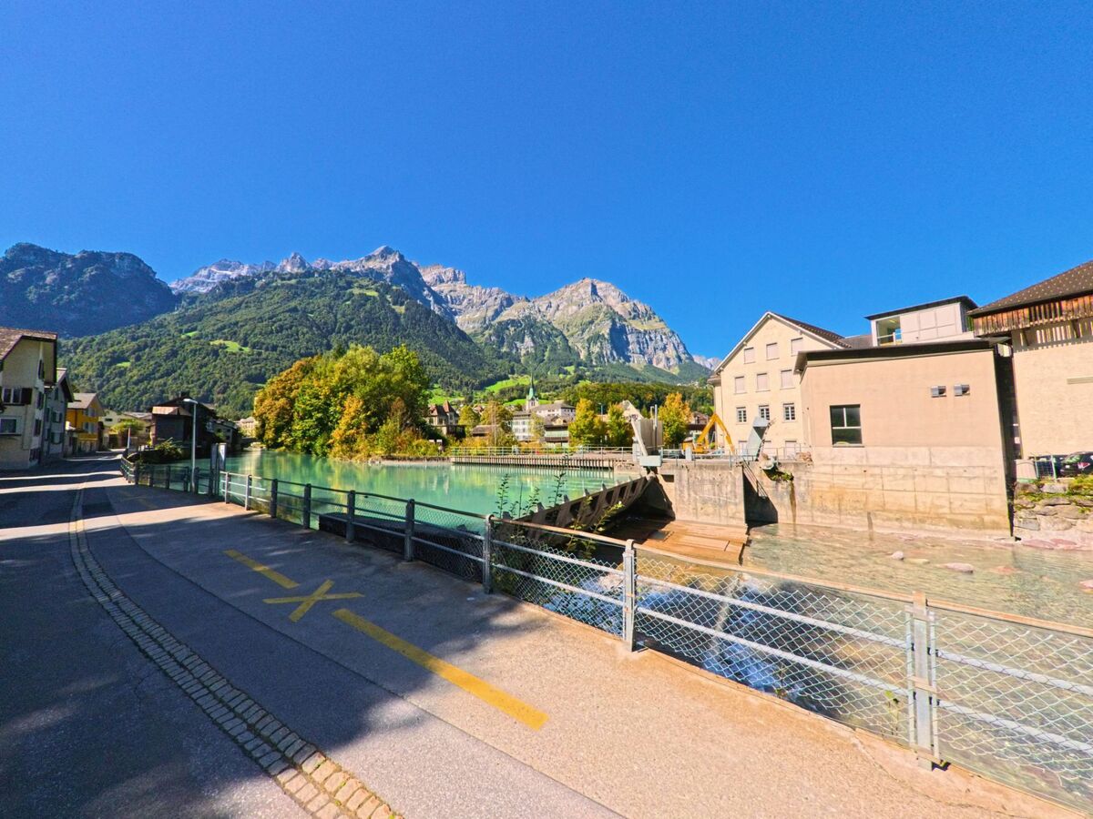 Ein klarer Fluss fliesst in Glarus_Sued (8762, Kanton Glarus) zwischen Wohnhaeusern und Industriegebaeuden, eingerahmt von steilen Alpenhaengen und blauem Himmel.