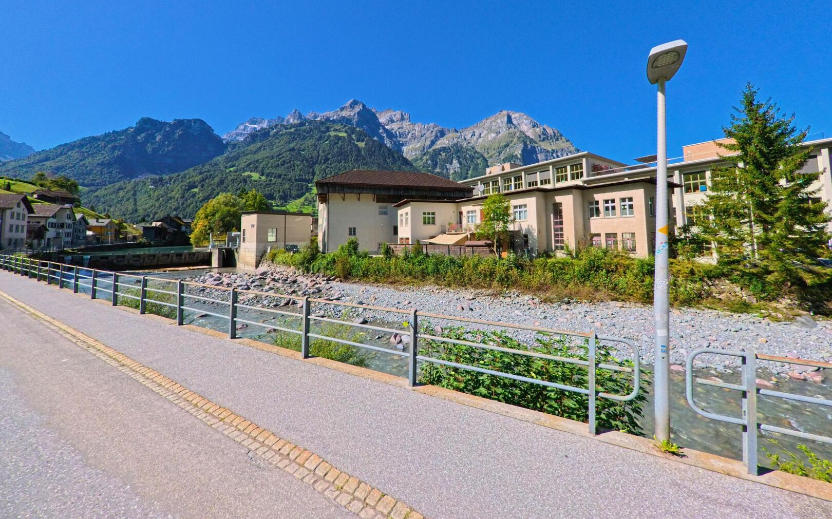 Ein Fussweg fuehrt in Glarus_Sued (8762, Kanton Glarus) entlang eines klaren Bergbachs, vorbei an Gebaeuden mit Blick auf die markanten Gipfel der umliegenden Alpen.