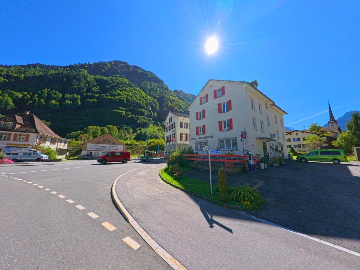 In Glarus_Sued (8783, Kanton Glarus) zeigt sich unter strahlender Sonne der Ortskern mit dem Hotel Eidgenossen und Blick auf die bewaldeten Hänge der umgebenden Berge.