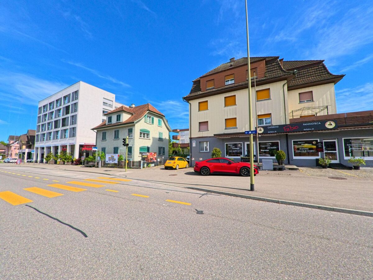 Entlang einer belebten Strasse in Glattbrugg (8152, Kanton Zürich) stehen ein modernes weisses Gebäude und ältere Wohnhäuser, vor denen mehrere Fahrzeuge parkiert sind.