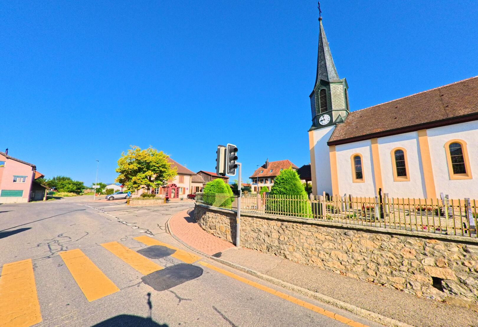 An einer Kreuzung in Gletterens (1544, Kanton Freiburg) steht die Dorfkirche mit ihrem markanten Turm neben einem Fussgaengerstreifen und einer Verkehrsampel.