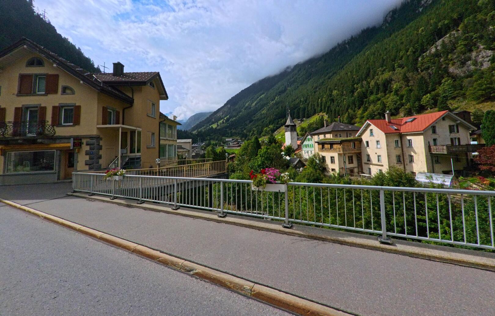 Von einer Bruecke in Göschenen (6487, Kanton Uri) blickt man auf Wohnhaeuser, die Kirche und die bewaldeten Berghaenge im Hintergrund.
