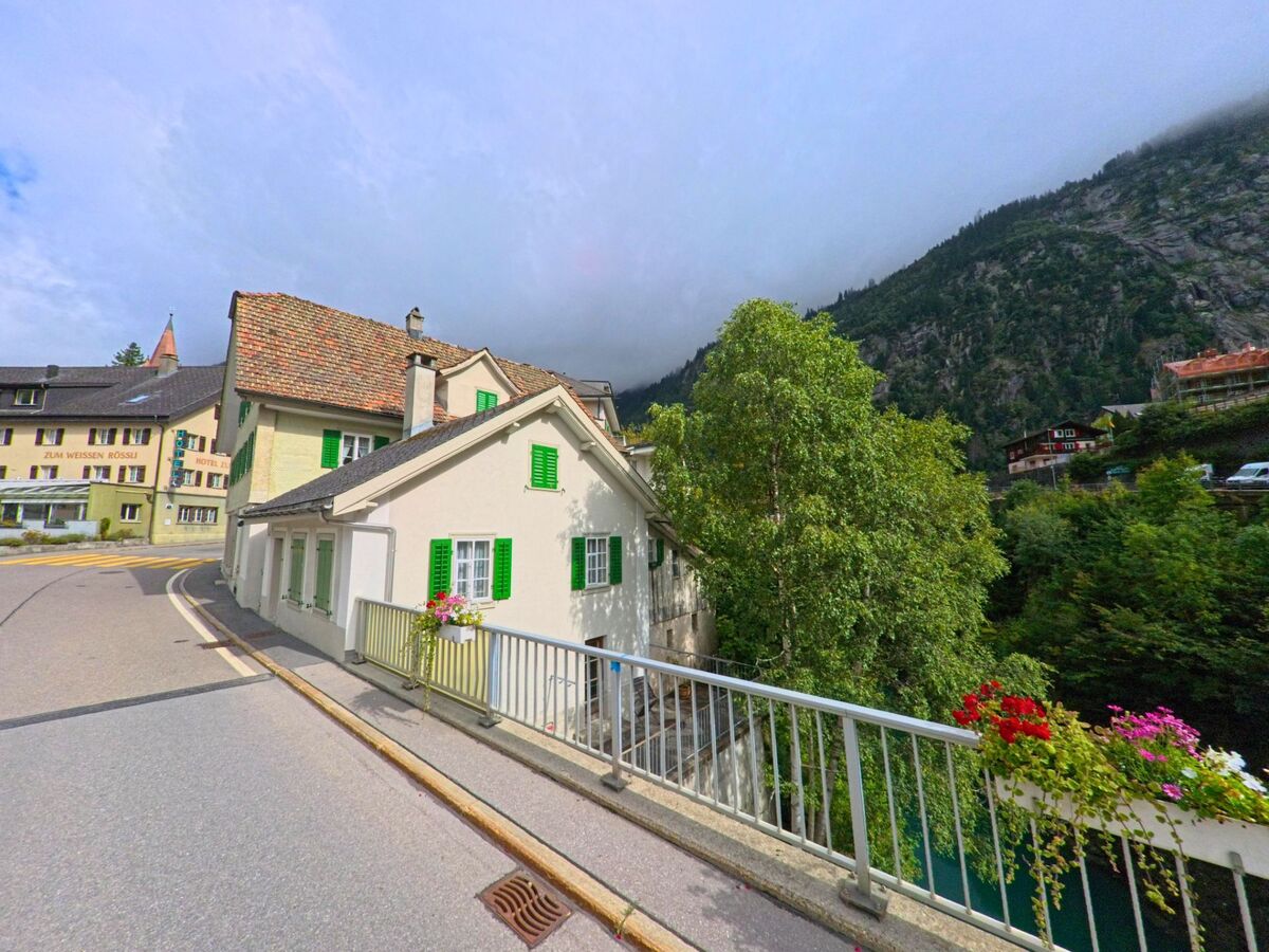 Ein Haus mit gruenen Fensterlaeden steht in Göschenen (6487, Kanton Uri) an einer Strasse, waehrend im Hintergrund die Berghaenge von Nebel umhüllt sind.