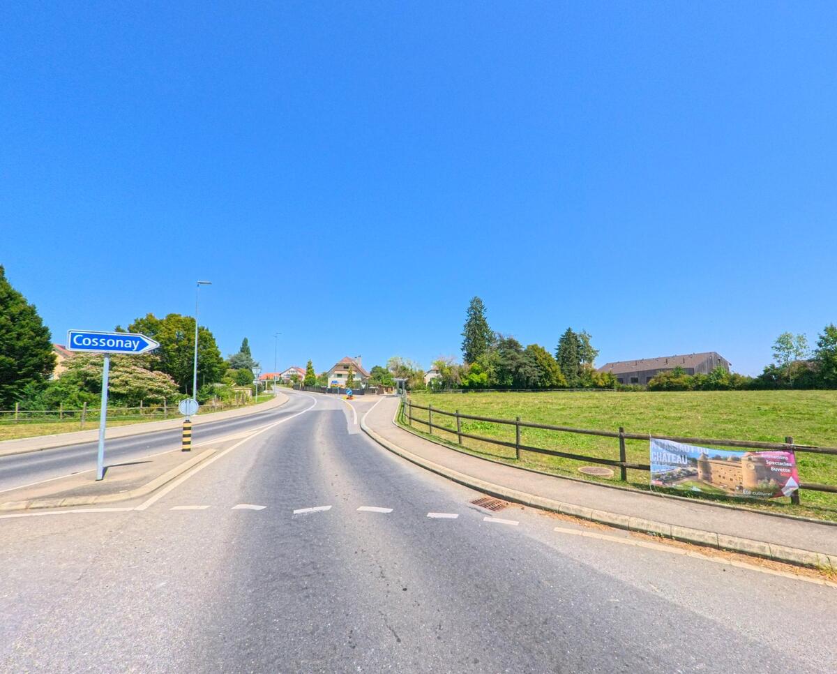 Von der Hauptstrasse in Gollion (1124, Kanton Waadt) fuehrt der Weg Richtung Cossonay, gesaeumt von Wiesen und einem Werbebanner am Strassenrand.