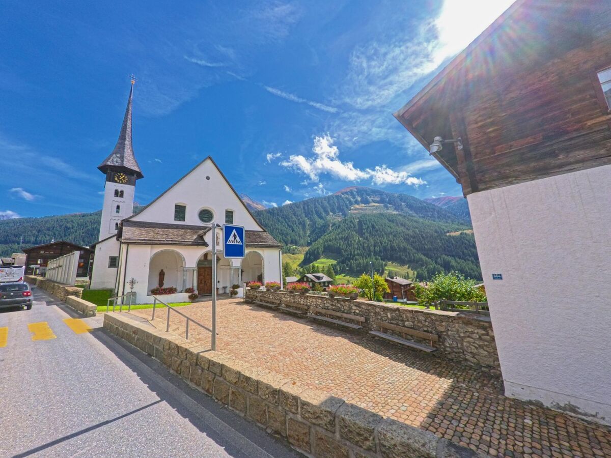 Die Kirche mit hohem Turm und Spitzdach in Goms (3985, Kanton Wallis) steht am Strassenrand vor bewaldeten Berghaengen.
