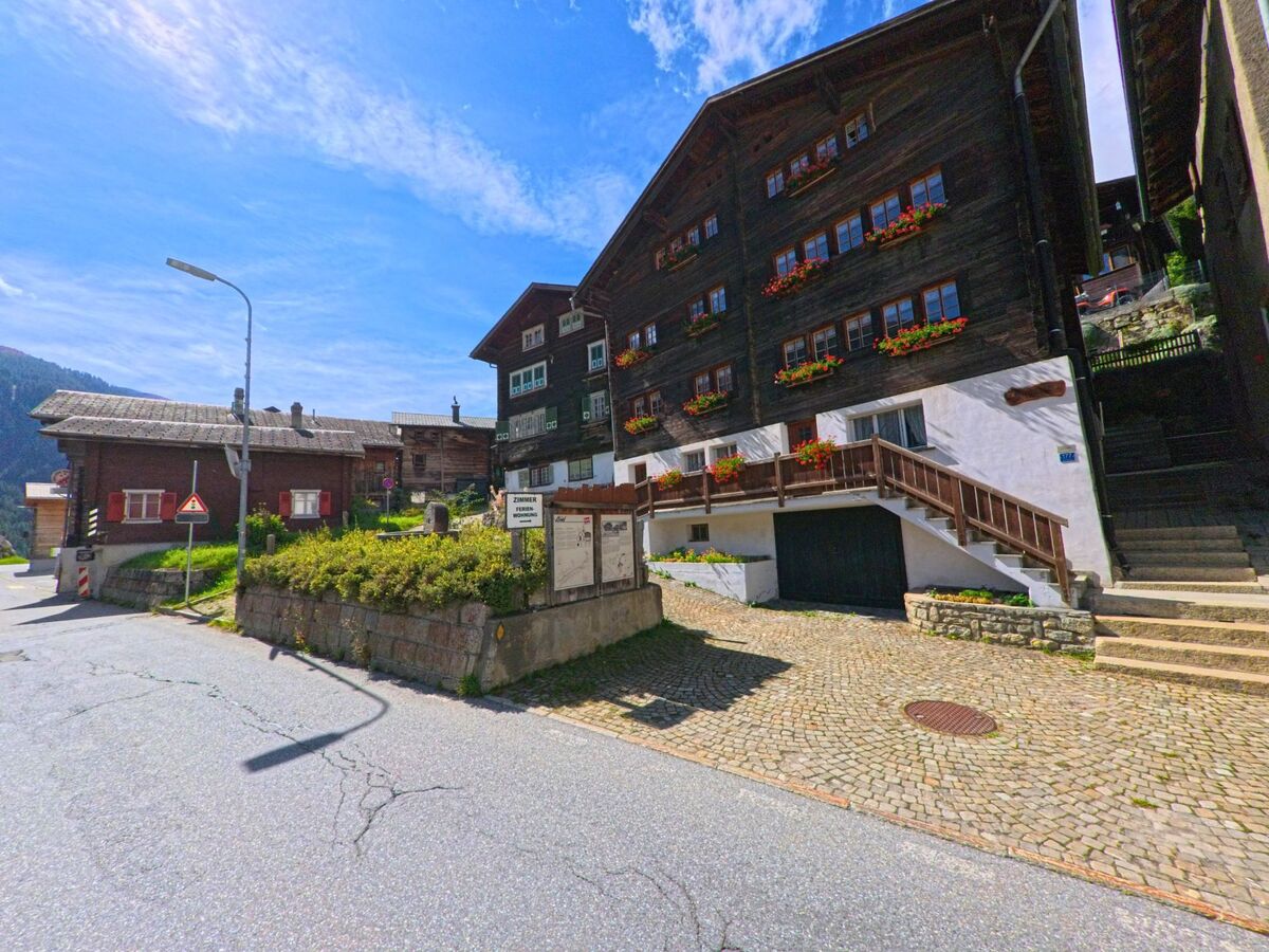 Traditionelle Holzhaeuser mit Blumenkisten saeumen in Goms (3989, Kanton Wallis) eine gepflasterte Strasse im Dorfzentrum.