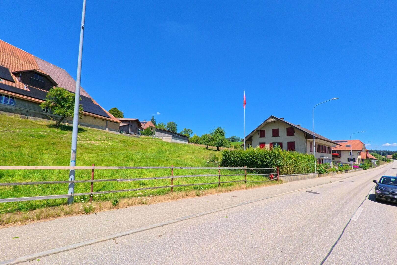 An der Hauptstrasse in Gondiswil (4955, Kanton Bern) reihen sich Wohnhaeuser und Bauerngebaeude mit Solarpanelen entlang eines gruenen Hangs unter blauem Himmel.