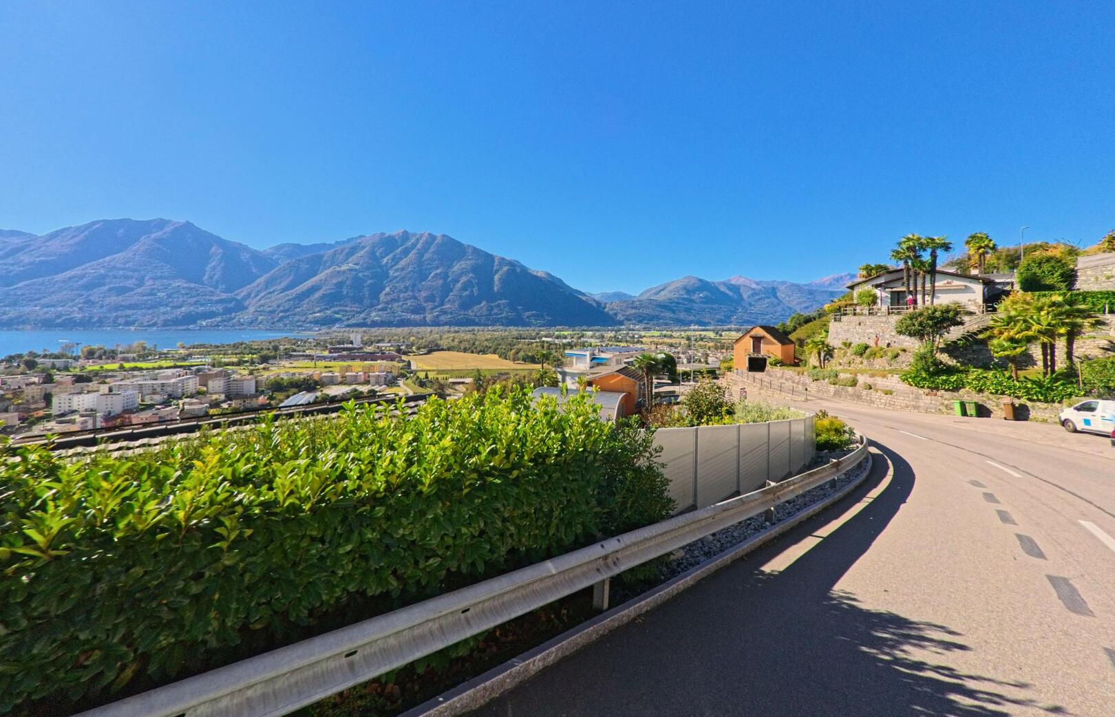 Von einer ansteigenden Strasse in Gordola (6596, Kanton Tessin) öffnet sich der Blick über die Häuser des Ortes hin zum Lago Maggiore und den umliegenden Bergen.