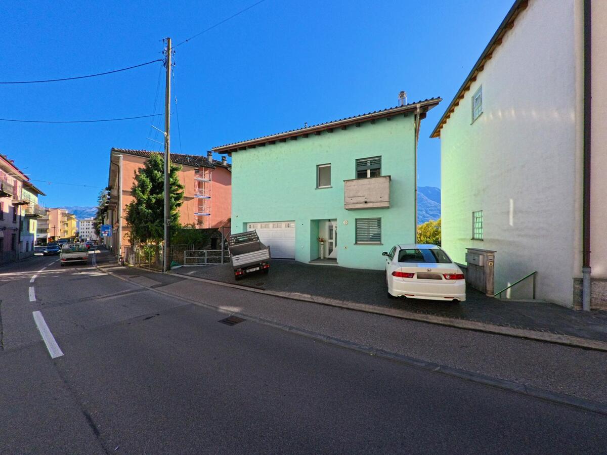 An einer Wohnstrasse in Gordola (6596, Kanton Tessin) stehen farbige Häuser mit Balkonen und geparkten Autos unter blauem Himmel.