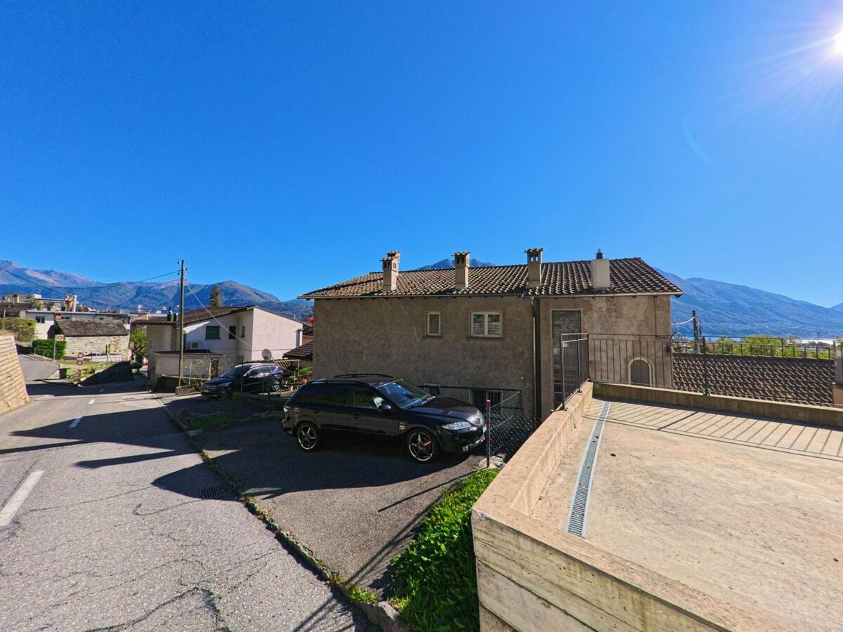 In einem Wohnquartier von Gordola (6596, Kanton Tessin) stehen Häuser mit Ziegeldächern und Blick auf die umliegenden Berge unter klarem Himmel.