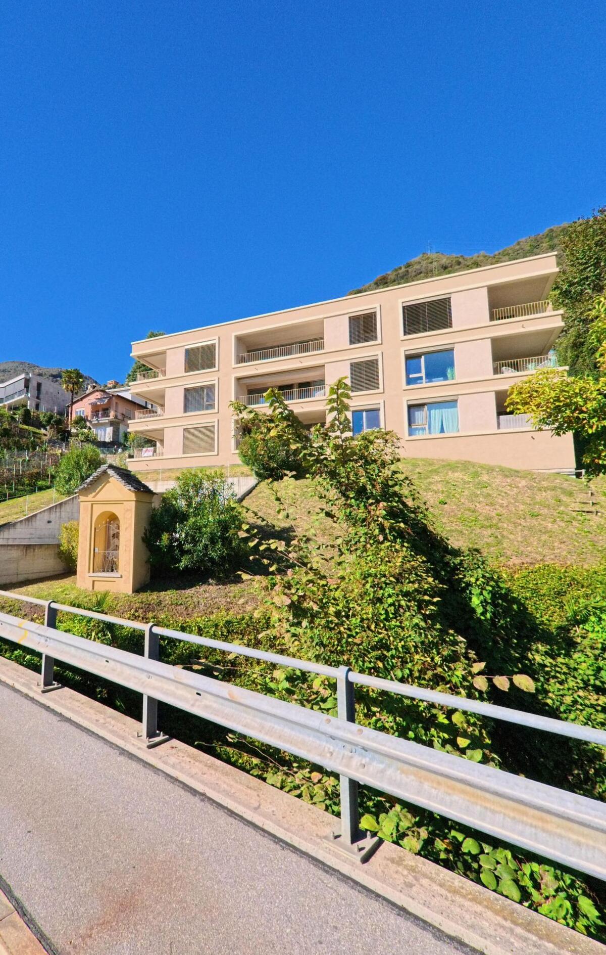 Ein modernes Mehrfamilienhaus in Gordola (6596, Kanton Tessin) steht an einem sonnigen Hang oberhalb einer kleinen Kapelle.