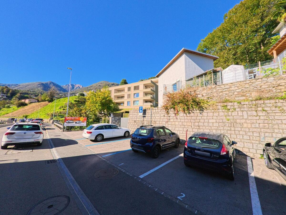 Ein Parkplatz in Gordola (6596, Kanton Tessin) liegt unterhalb moderner Wohnhäuser mit Blick auf die grüne Hanglandschaft und Berge.