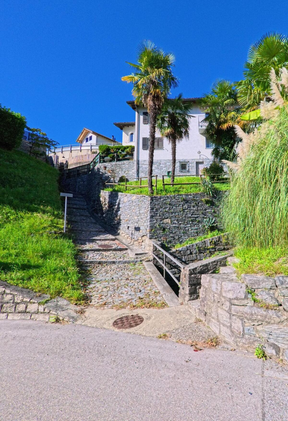 Ein schmaler Pflasterweg in Gordola (6596, Kanton Tessin) führt zwischen Steinmauern und Palmen zu einem Haus am Hang hinauf.