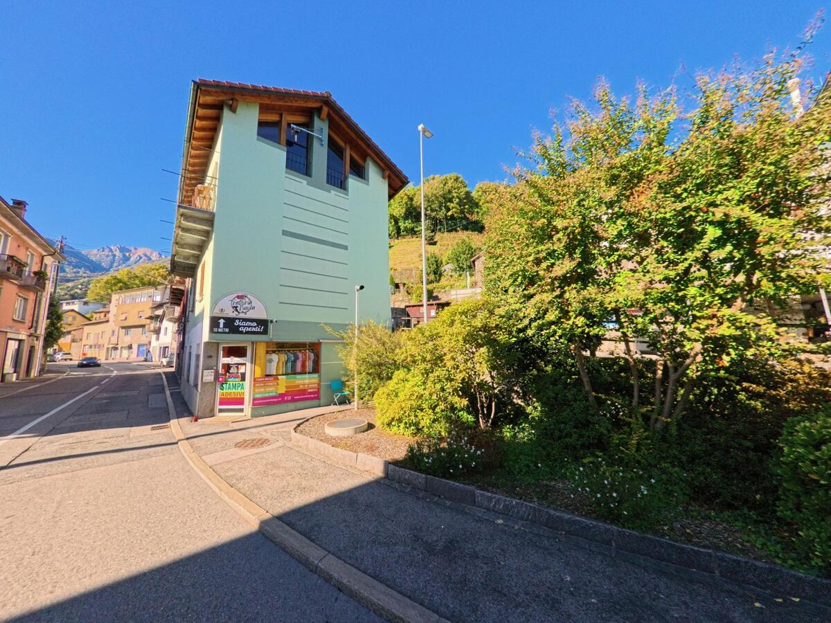 Ein schmaler, grüner Bau mit einem kleinen Laden steht an einer Kreuzung in Gordola (6596, Kanton Tessin) unter klarem Himmel.