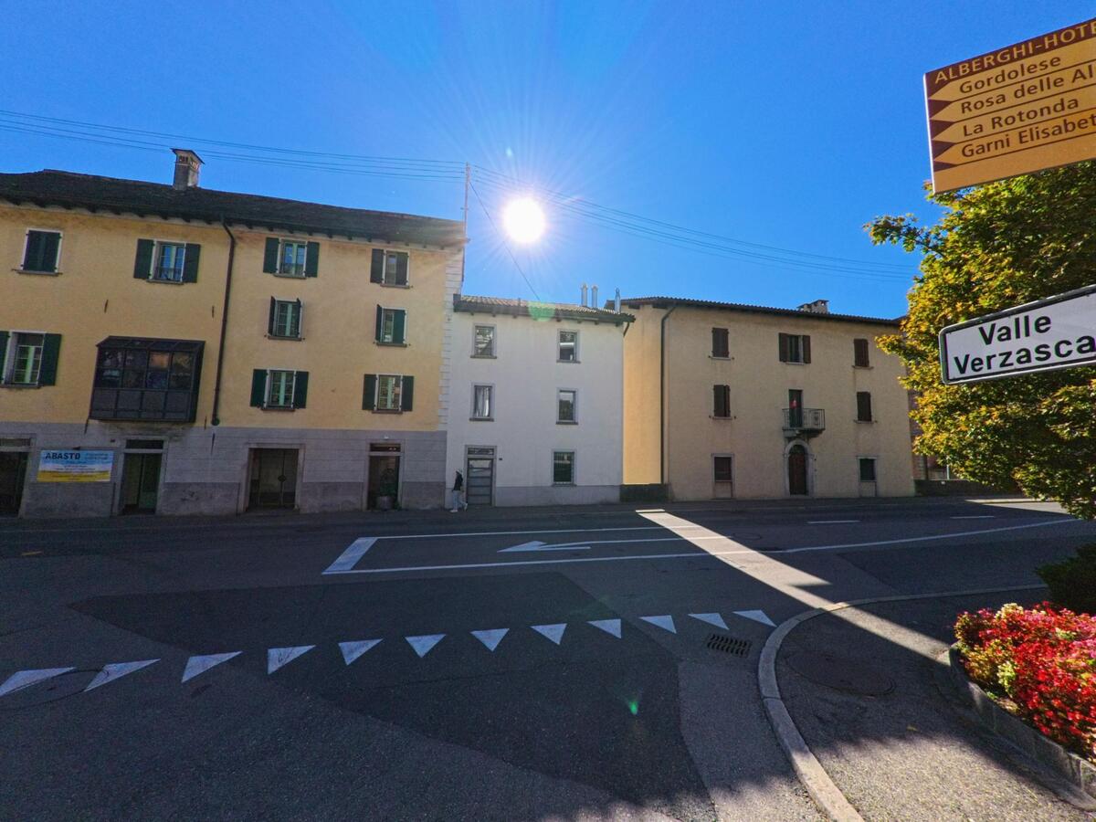 An einer Strassenkreuzung in Gordola (6596, Kanton Tessin) weisen Schilder den Weg ins Valle Verzasca, während historische Häuser in der Sonne stehen.