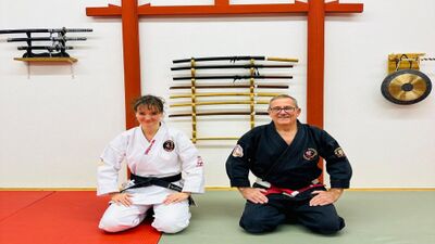 In Liestal (4410, Kanton Baselland) knien zwei Kampfsporttreibende im Dojo vor einer Wand mit Trainingswaffen und zeigen ruhige Konzentration in traditioneller Kleidung.