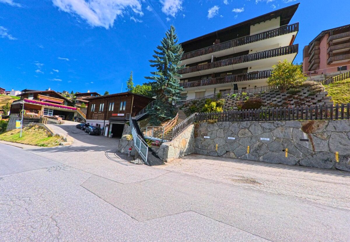 An einem Hang in Grächen (3925, Kanton Wallis) stehen ein mehrstöckiges Wohnhaus mit Balkonen und mehrere kleinere Holzbauten, die von einer steilen Strasse erschlossen werden.