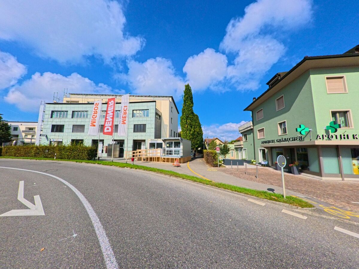 An einer Strassenkurve in Graenichen (5722, Kanton Aargau) stehen moderne Gebaeude mit einer Apotheke und einem Buerohaus unter blauem Himmel.