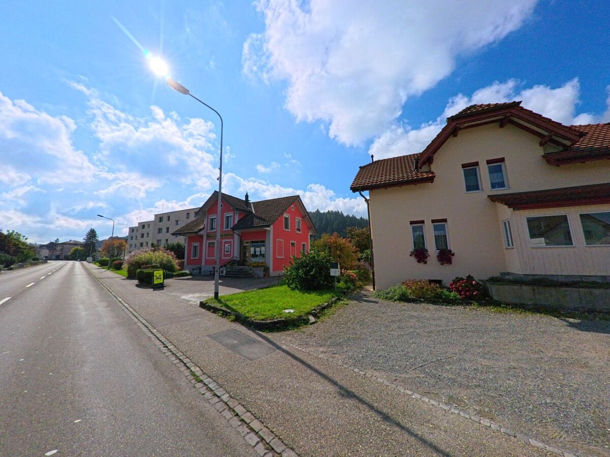 Entlang einer sonnigen Strasse in Graenichen (5722, Kanton Aargau) stehen farbenfrohe Wohnhaeuser, darunter ein rotes Haus mit gepflegtem Vorgarten.