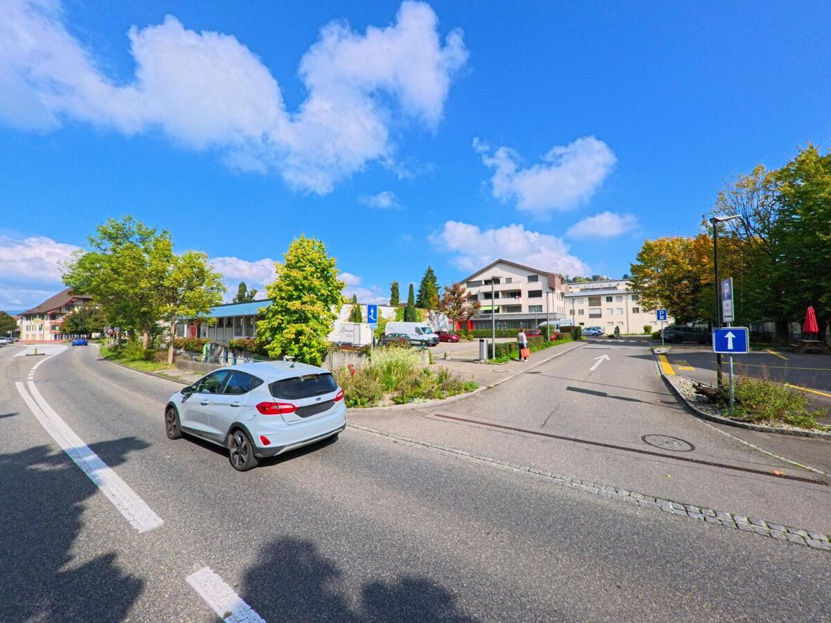 Ein Auto faehrt auf einer Hauptstrasse in Graenichen (5722, Kanton Aargau) vorbei an modernen Wohngebaeuden und Parkplaetzen bei sonnigem Wetter.