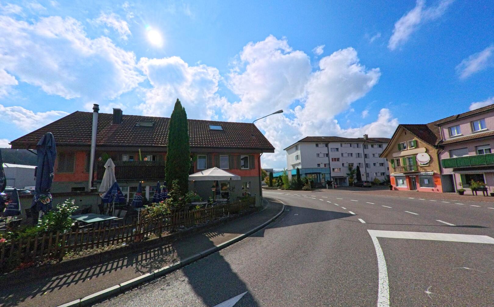 An einer Kreuzung in Graenichen (5722, Kanton Aargau) stehen mehrere Wohnhaeuser mit Balkonen und farbigen Fassaden unter einem wolkigen Himmel in der Sonne.