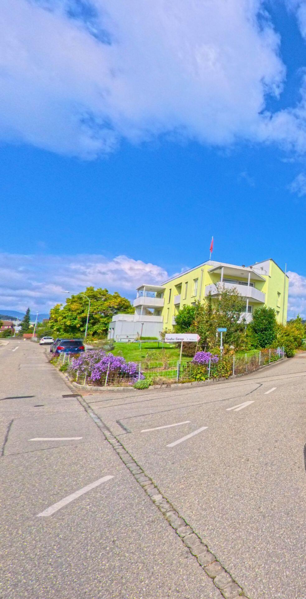 Ein modernes, hellgruenses Mehrfamilienhaus mit Balkonen und Garten befindet sich an einer Strassenecke in Graenichen (5722, Kanton Aargau) unter blauem Himmel.