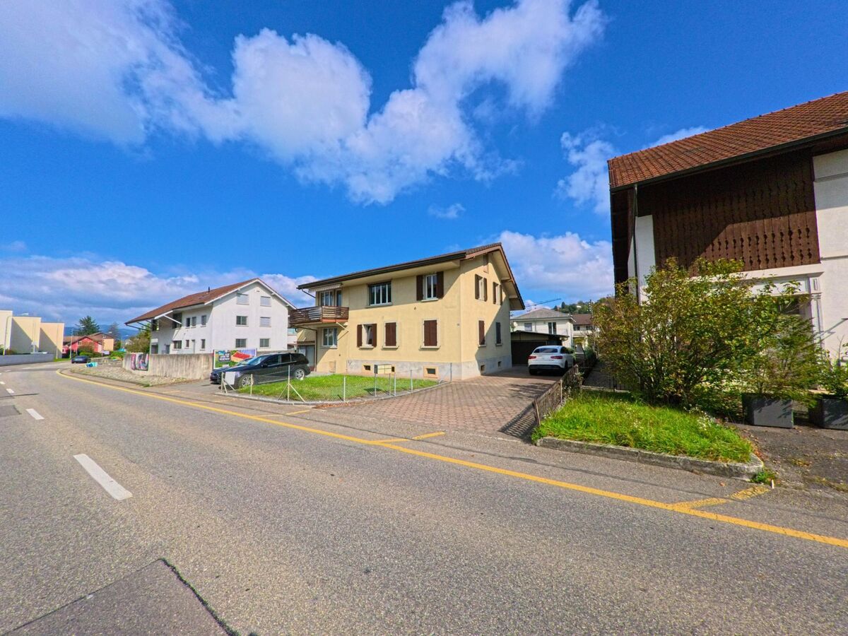 Mehrere Wohnhaeuser mit Einfahrten und Gaerten saeumen eine leicht gekruemmte Strasse in Graenichen (5722, Kanton Aargau) unter blauem Himmel.