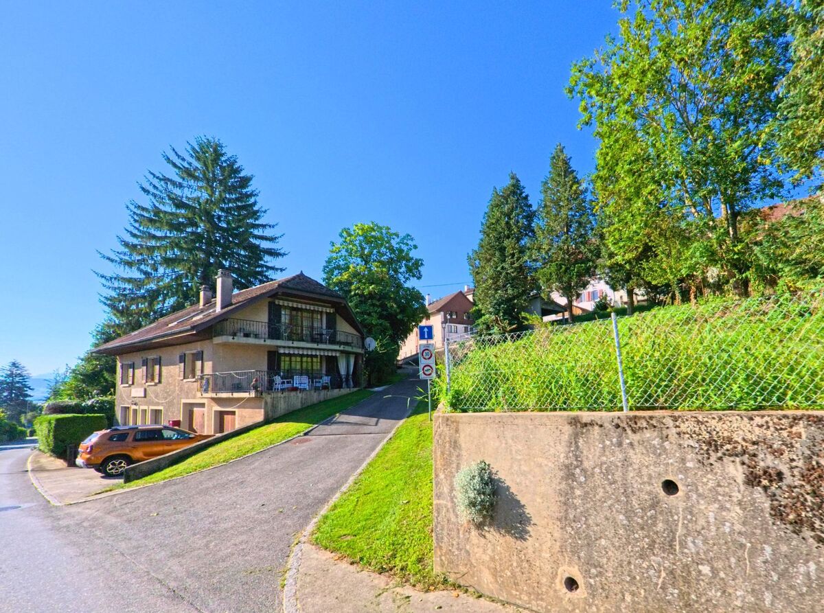Ein Einfamilienhaus mit Balkon steht in Grandcour (1543, Kanton Waadt) an einer steilen Auffahrt, umgeben von grossen Bäumen und gepflegten Grünflächen.