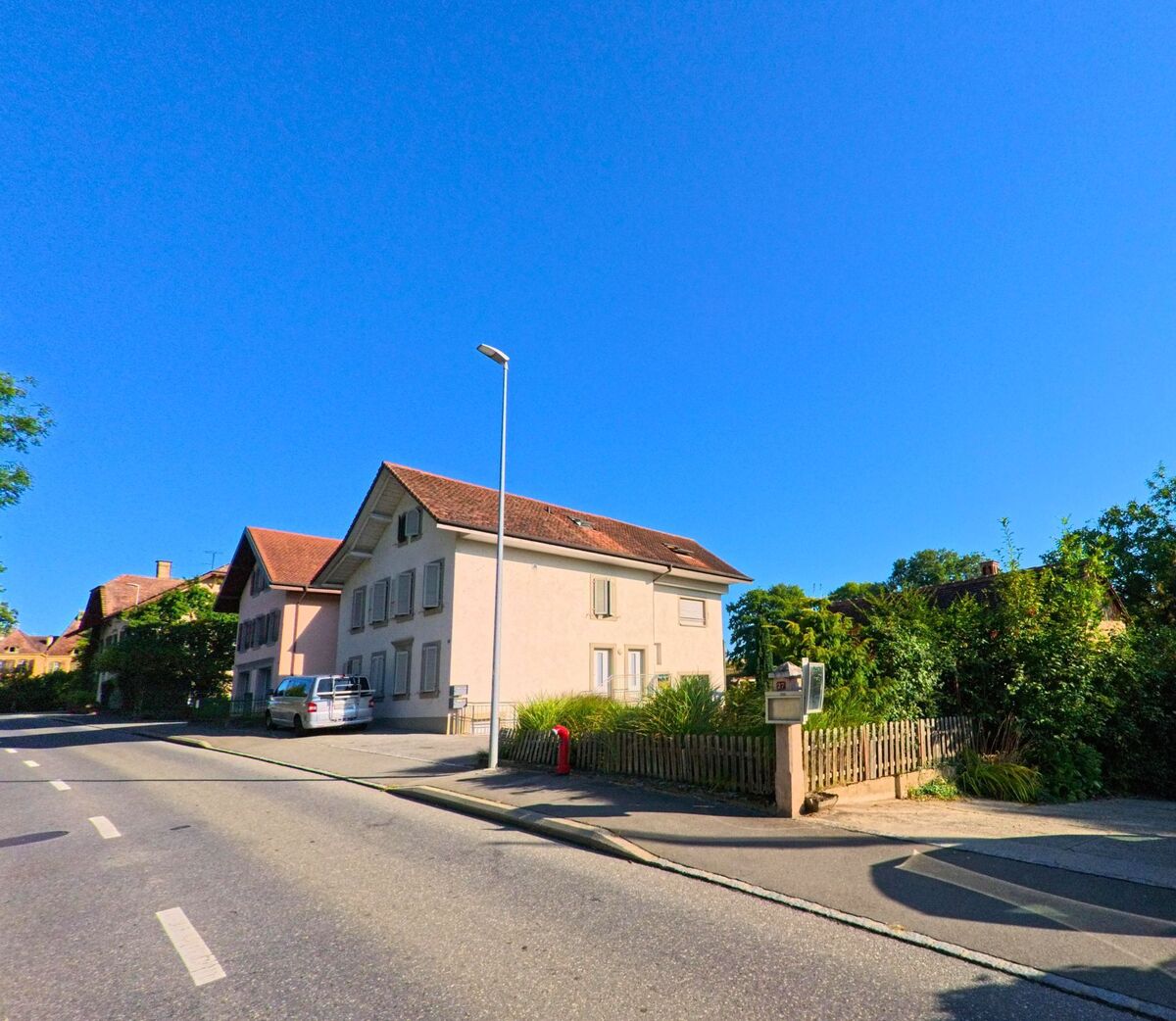 An einer Hauptstrasse in Grandcour (1543, Kanton Waadt) steht ein helles Wohnhaus mit geschlossenen Fensterläden und kleinem Vorgarten hinter einem Holzzaun.