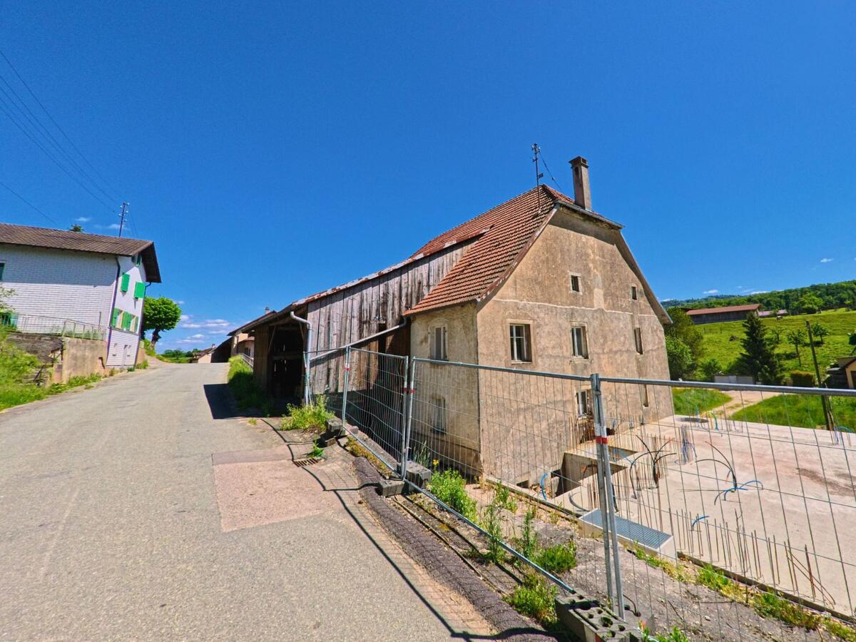Ein altes Haus mit verwitterter Fassade in Grandfontaine (2908, Kanton Jura) steht an einer leicht ansteigenden Strasse neben einer Baustelle und offenen Feldern.