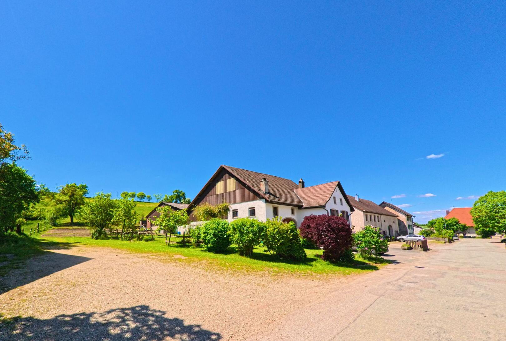 In Grandfontaine (2908, Kanton Jura) säumen gepflegte Bauernhäuser mit Gärten eine ruhige Dorfstrasse am Rand grüner Hügel.