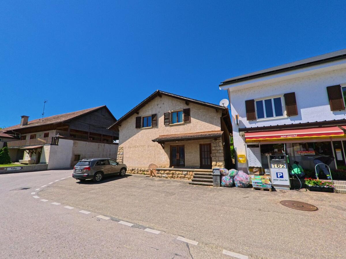Im Zentrum von Grandfontaine (2908, Kanton Jura) stehen ein kleiner Dorfladen mit Tankstelle, ein Wohnhaus und ein Bauernhof nebeneinander unter strahlend blauem Himmel.