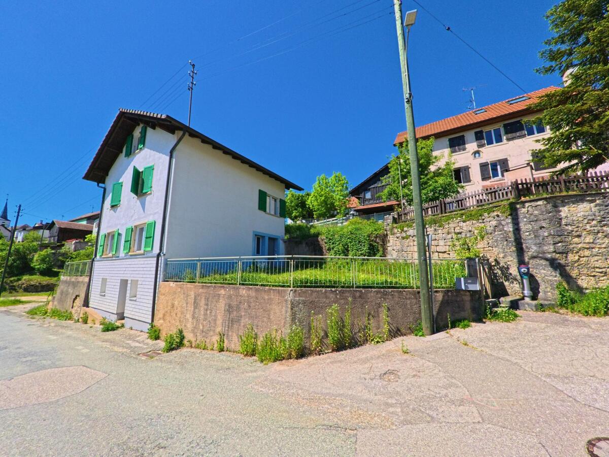Ein Haus mit grünen Fensterläden in Grandfontaine (2908, Kanton Jura) steht erhöht auf einer Mauerterrasse unter blauem Himmel neben einer Ladestation.