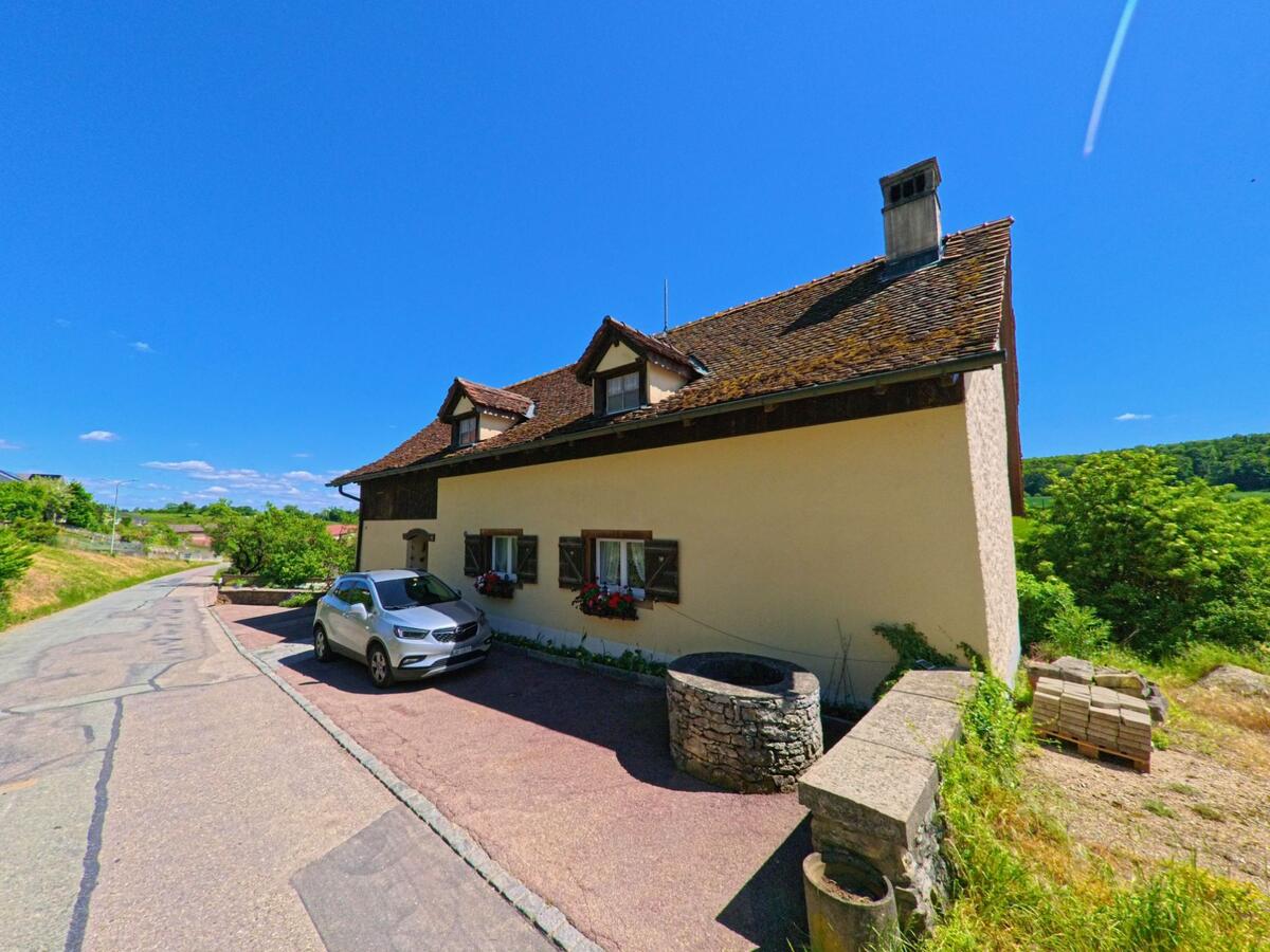 In Grandfontaine (2908, Kanton Jura) steht ein charmantes Haus mit blumengeschmückten Fensterläden, davor ein alter Steinbrunnen und ein geparktes Auto.
