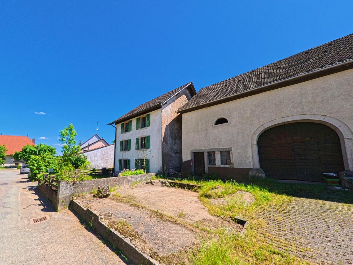 In Grandfontaine (2908, Kanton Jura) stehen ein Wohnhaus mit grünen Fensterläden und eine Scheune mit grossem Holztor nebeneinander an einer ruhigen Strasse.