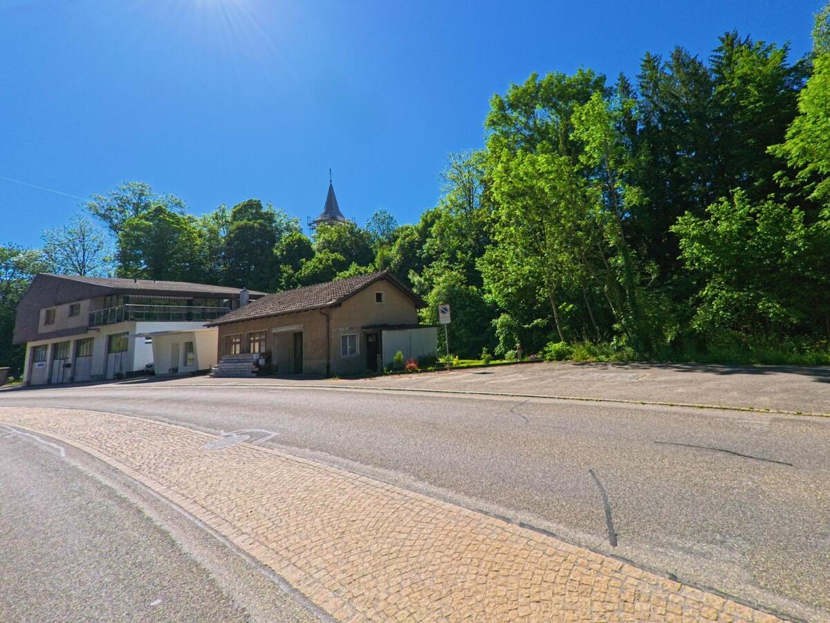 Hinter einem kleinen Gebäudekomplex an einer Straße in Grandval (2745, Kanton Bern) ragt der Kirchturm über die Baumkronen eines angrenzenden Waldes hinaus.