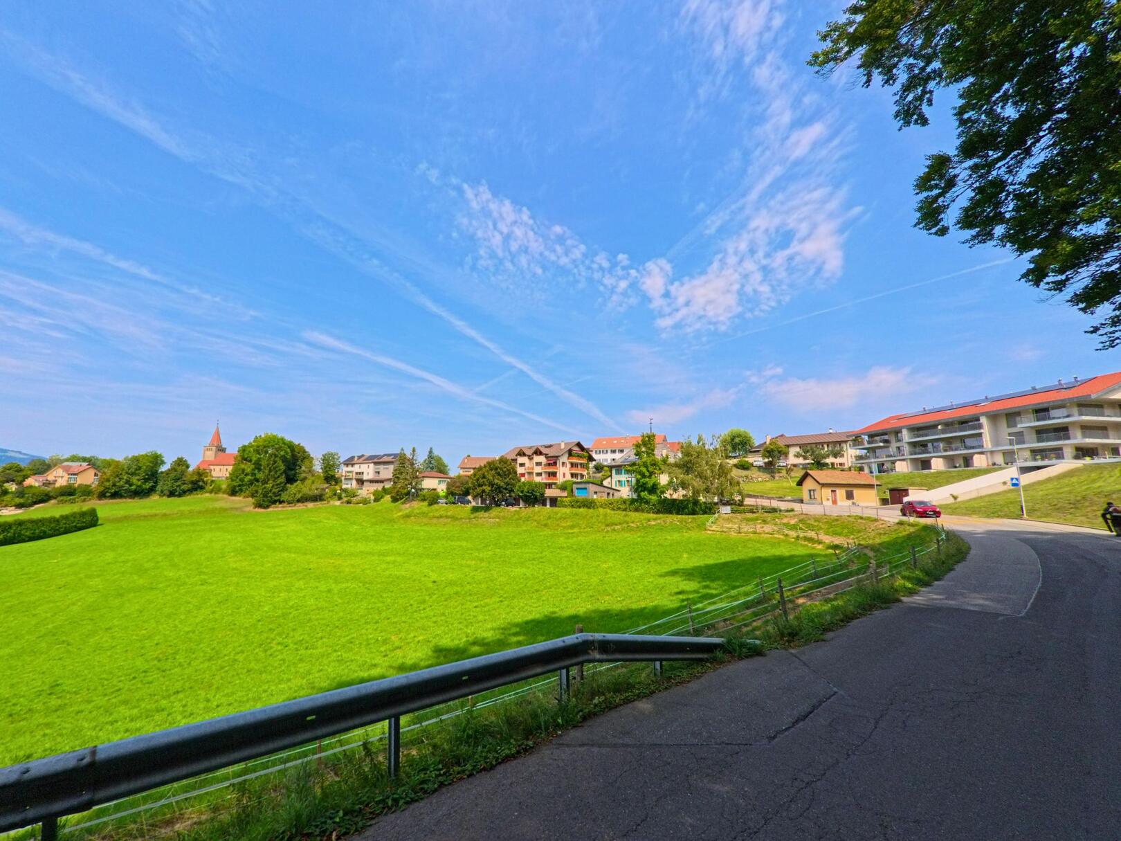 Von der Strasse aus bietet sich in Granges_(Veveyse) (1614, Kanton Freiburg) ein weiter Blick auf grüne Wiesen und Wohnhäuser mit rotem Kirchendach.