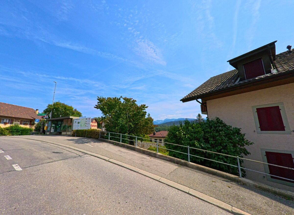 An einer Strasse in Granges_(Veveyse) (1614, Kanton Freiburg) befindet sich eine Bushaltestelle mit Wartehaus und Schildern, im Hintergrund sind Berge sichtbar.
