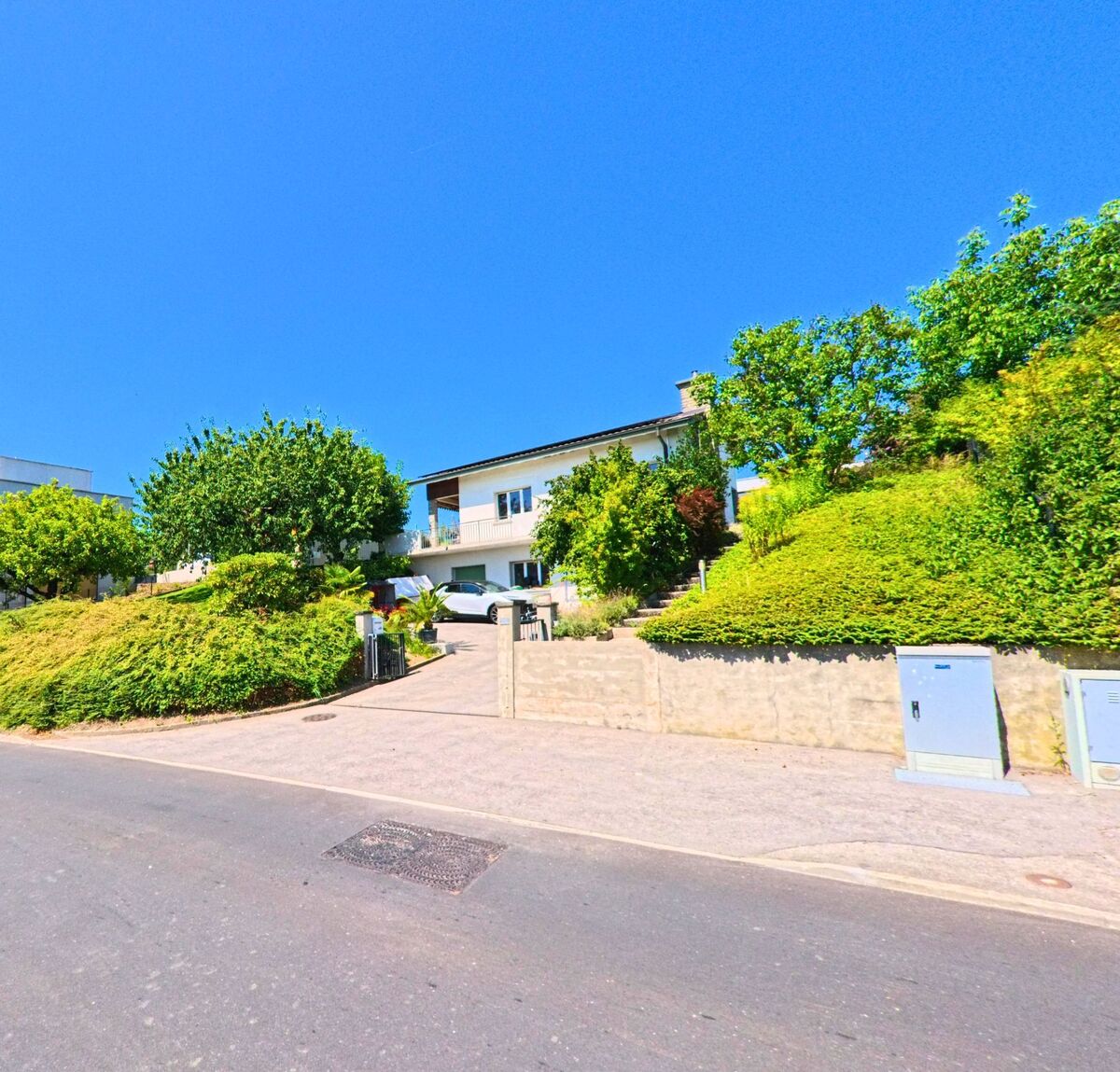 Ein Einfamilienhaus mit Balkon und Auffahrt liegt in Granges-Paccot (1763, Kanton Freiburg) auf einer von Baeumen und Straeuchern umgebenen Anhöhe.