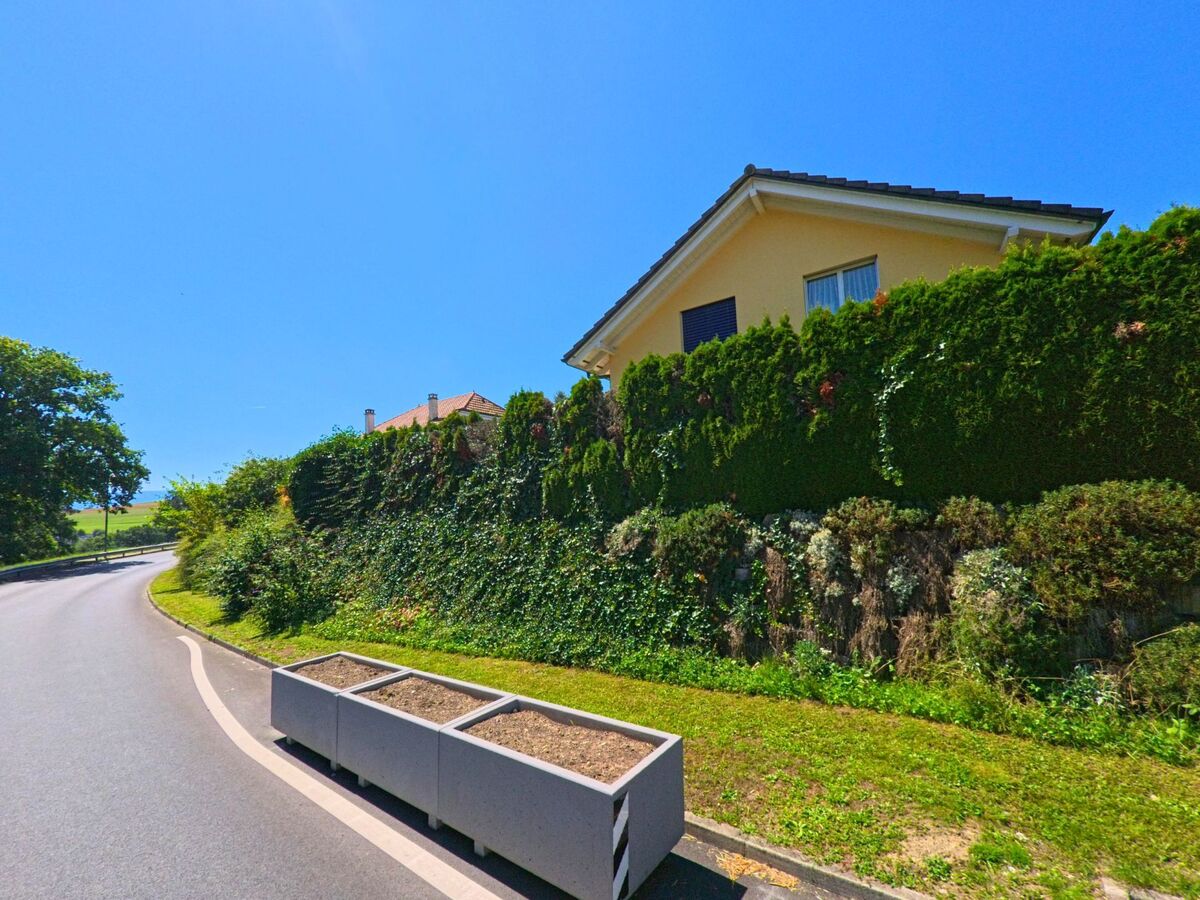 An einer leicht geschwungenen Strasse in Granges-Paccot (1763, Kanton Freiburg) steht ein gelbes Wohnhaus hinter einer hohen, dichten Hecke.