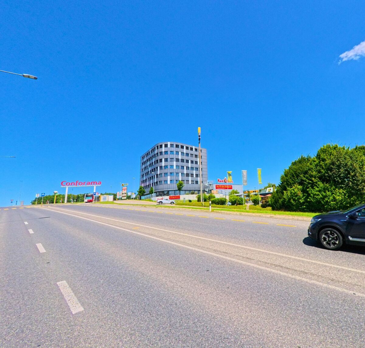 Entlang einer breiten Strasse in Granges-Paccot (1763, Kanton Freiburg) stehen ein modernes Hochhaus und mehrere Geschaefte wie Conforama und Yendi.