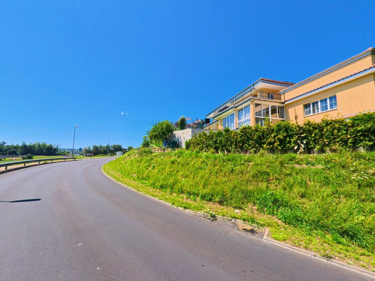 Eine leicht ansteigende Strasse fuehrt in Granges-Paccot (1763, Kanton Freiburg) an einem gelben Gebaeude mit grossen Fenstern und Terrasse vorbei.