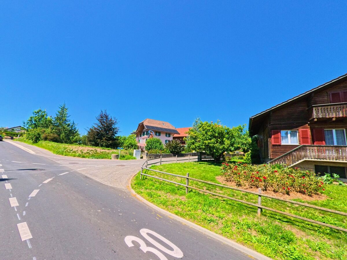 Zwei Wohnhaeuser, eines in Holzbauweise, stehen in Granges-Paccot (1763, Kanton Freiburg) an einer verkehrsberuhigten Strasse mit Tempo-30-Markierung.