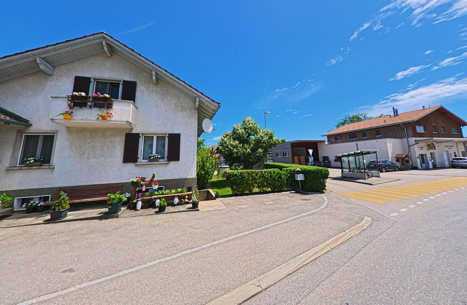 In Grangettes (1686, Kanton Freiburg) steht ein Wohnhaus mit Blumenschmuck an Fenstern und Balkon neben einer Bushaltestelle, die an einer verkehrsberuhigten Strasse liegt.