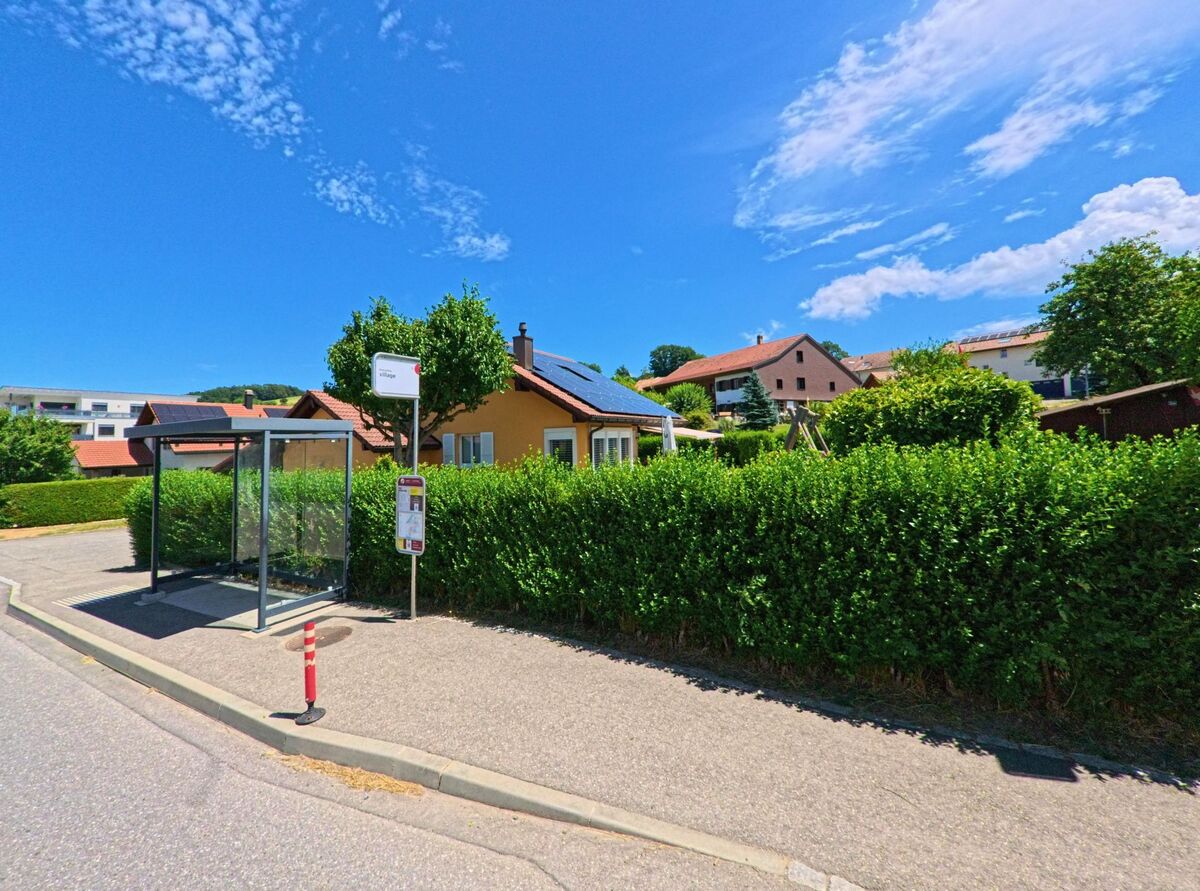 In einem Wohnquartier von Grangettes (1686, Kanton Freiburg) befindet sich eine kleine Bushaltestelle vor einem Haus mit Solarpanels und gepflegter Hecke.