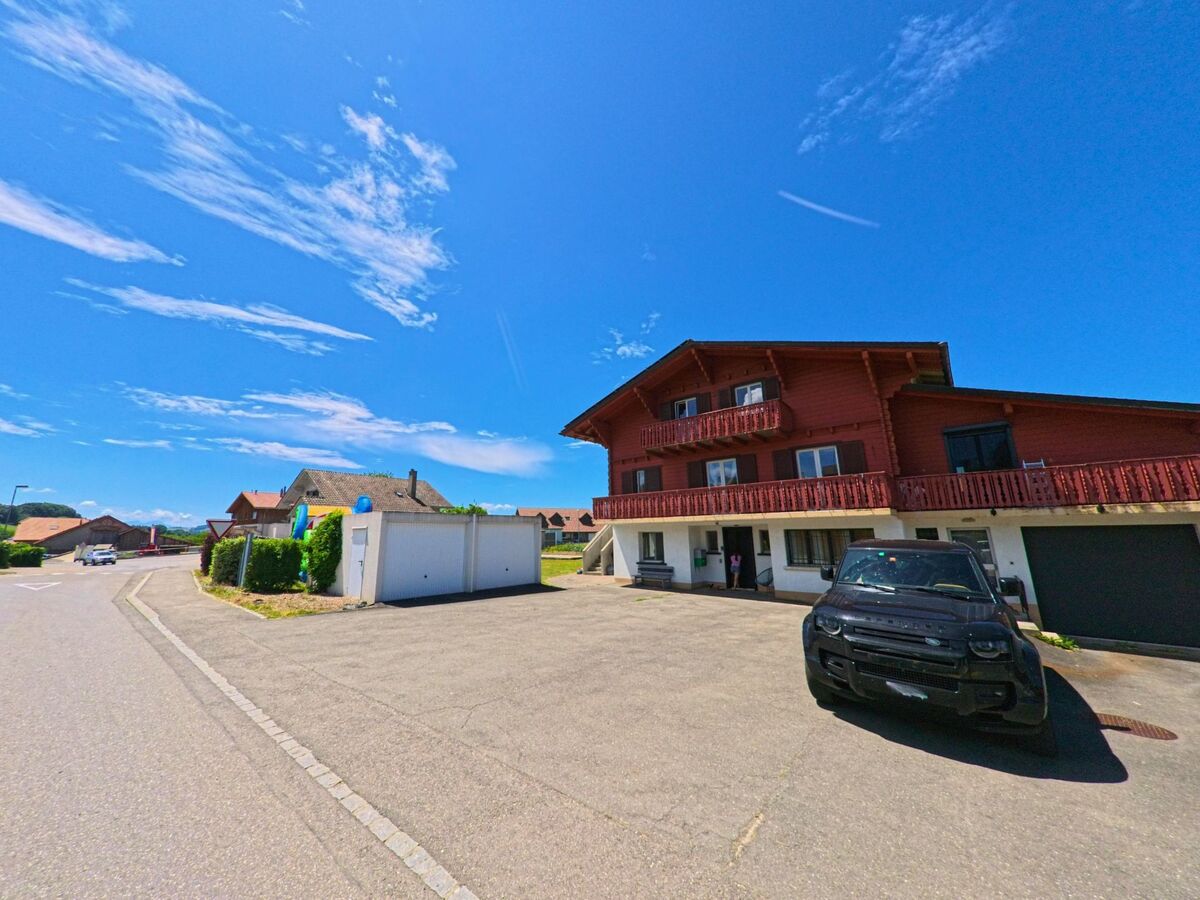 In Grangettes (1686, Kanton Freiburg) steht ein modernes Chalet mit roten Holzbalkonen an einer ruhigen Wohnstrasse, vor dem ein schwarzes Fahrzeug parkt.